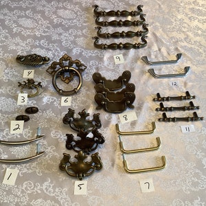 May include: A selection of antique cabinet hardware, including drawer pulls and knobs. The hardware features various designs, including ornate brass handles, ring pulls, and decorative knobs. The pieces are in shades of gold, brass, and silver.