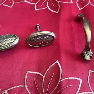 Può includere: Tre pomelli per armadi in ottone antico. Due sono di forma ovale con un design intrecciato e uno è una maniglia curva con un'estremità decorativa.