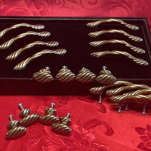 Può includere: Una collezione di 18 pomelli e maniglie per armadi dorati con un design a corda. I pomelli sono piccoli e rotondi, mentre le maniglie sono più lunghe e curve. I pomelli e le maniglie sono disposti su una superficie di velluto nero.
