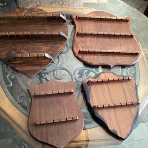 Peut inclure: Quatre présentoirs à cuillères en bois de différentes formes de boucliers et de cœurs. Chaque présentoir comporte trois rangées de petits chevilles en bois pour tenir les cuillères. Le bois est teinté brun foncé, et les présentoirs sont exposés sur une table en verre.