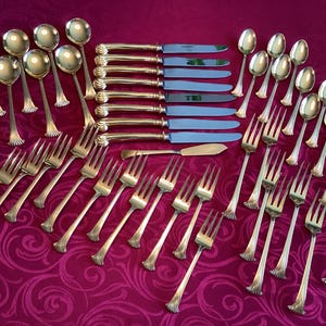 May include: A set of vintage silverware, including spoons, forks, knives, and a butter knife, arranged on a patterned burgundy surface. The silverware has a silver finish with ornate handles and shiny blades.