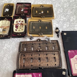 May include: An assortment of decorative wall plate switch covers in various styles and colours. Some are plain white, others have a floral design, and some have a gold metallic finish. The plates are packaged in clear plastic bags.