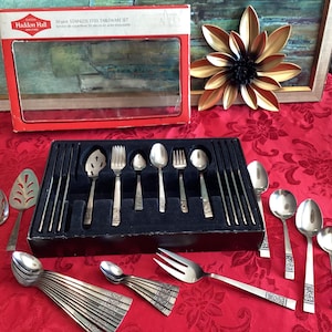 Puede incluir: Un juego de cubiertos de acero inoxidable vintage, que incluye cucharas, tenedores y utensilios para servir, dispuestos en una caja negra con una etiqueta roja y blanca. El juego se exhibe sobre un mantel rojo floral.