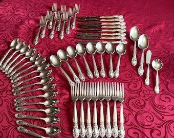 May include: A set of silverware with ornate handles, including forks, spoons, and knives. The silverware is arranged on a red tablecloth with a floral pattern.