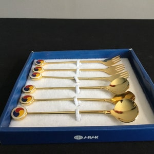 May include: A set of six gold-colored spoons and forks in a blue box. The spoons and forks have a white and red design on the handle. The box has the word "ARAK" printed on the bottom.