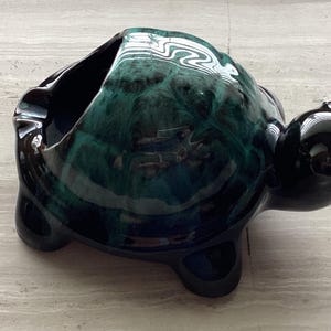 Może przedstawiać: Ceramiczna popielniczka w kształcie żółwia. Popielniczka jest szkliwiona w odcieniach ciemnego turkusu i czerni, z błyszczącym wykończeniem. Żółw ma duży otwór na popiół i gładki, zaokrąglony pancerz.