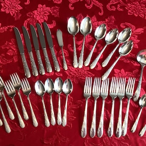 May include: An assortment of silver-coloured cutlery, including forks, knives, spoons, and a ladle, displayed on a red floral tablecloth. The silverware features decorative floral patterns on the handles, suitable for a formal dining occasion.