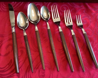 Op de afbeelding: Een set van zes roestvrijstalen bestekdelen met donkerbruine houten handvatten. De collectie bevat een tafelmes, twee lepels van verschillende groottes en drie vorken van verschillende groottes, allemaal op een rij op een rode ondergrond.