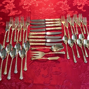 May include: A set of gold-plated cutlery, including forks, knives, and spoons, laid out on a red tablecloth. The cutlery has an ornate design.