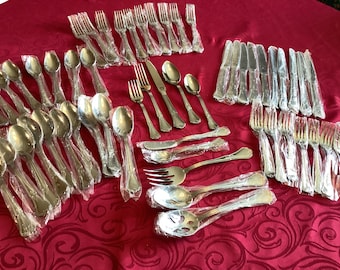 May include: A set of silverware in individual plastic packaging. The set includes forks, knives, spoons, and serving utensils. The silverware is arranged on a red tablecloth with a floral pattern.