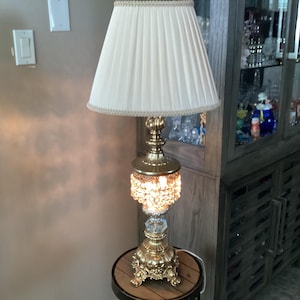 May include: A gold table lamp with a white lampshade and a crystal base. The lamp is on a wooden table with a brown surface.
