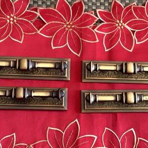 Peut inclure: Quatre poignées de porte de placard en laiton antique avec une base rectangulaire et un design décoratif. Les poignées ont une finition brossée et sont en bon état.