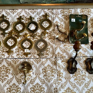 May include: A collection of brass-toned hardware including nine circular decorative plates with ornate cutouts, a shell-shaped hook, two double-pronged hooks with white tips on a card labeled 2, a single curved hook, and two wall-mounted hooks with wooden knobs.
