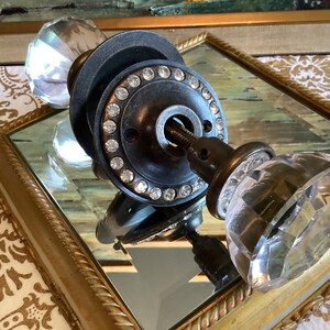 May include: A door knob set featuring two clear, multi-faceted glass knobs attached to a dark metal circular plate. The plate is embellished with a ring of small, clear, sparkling rhinestones around the center spindle and mounting holes.