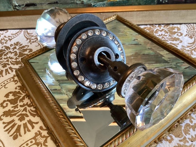 May include: A door knob set featuring two clear, multi-faceted glass knobs attached to a dark metal circular plate. The plate is embellished with a ring of small, clear, sparkling rhinestones around the center spindle and mounting holes.