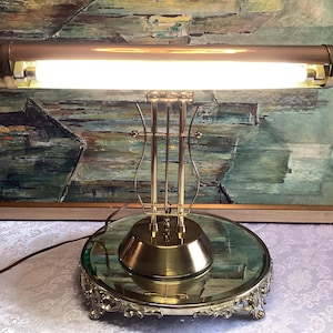 May include: A vintage brass desk lamp with a long, rectangular light fixture. The lamp has a round base with an ornate, decorative edge and a clear glass top. The lamp is on a table in front of a framed painting.
