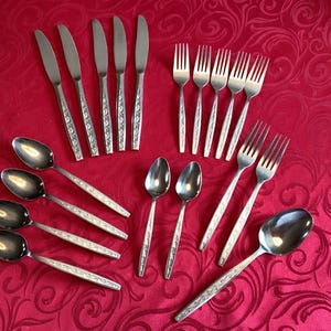 Puede incluir: Un juego de cubiertos de acero inoxidable, que incluye cucharas, tenedores y cuchillos, dispuestos sobre un mantel rojo estampado. Los cubiertos tienen mangos decorativos. La imagen muestra un juego de comedor completo.