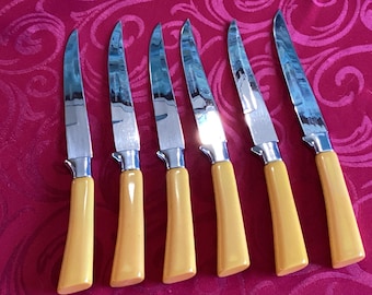 Op de afbeelding: Een set van zes steakmessen met gepolijste zilverkleurige metalen bladen en gladde, effen gele handvatten. Elk mes heeft een gebogen lemmet en een krop waar het blad het handvat ontmoet, gerangschikt in een rij op een rode ondergrond met patroon.