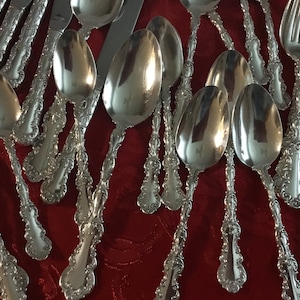 May include: A collection of silver-plated spoons and forks with ornate, decorative handles. The silverware is arranged on a red surface.