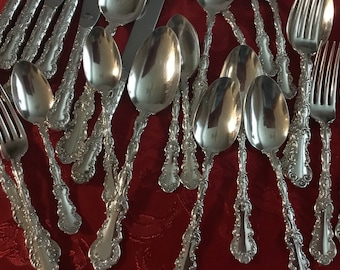 May include: A collection of silver-plated spoons and forks with ornate, decorative handles. The silverware is arranged on a red surface.