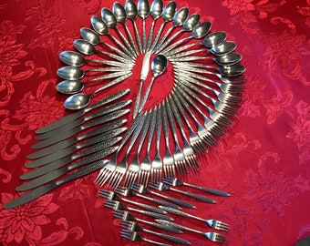 May include: A set of silverware arranged in a fan shape on a red tablecloth. The silverware includes spoons, forks, and knives, all with a decorative pattern.