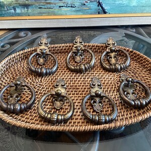 May include: Seven antique bronze door knockers with a floral design, presented on a woven wicker tray. Each knocker features a ring handle. The tray is displayed on a glass table.