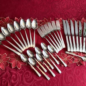 May include: A set of silverware with a silver finish and gold accents. The set includes spoons, forks, and knives. The silverware is arranged on a red tablecloth.