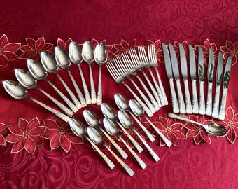 May include: A set of silverware with a silver finish and gold accents. The set includes spoons, forks, and knives. The silverware is arranged on a red tablecloth.