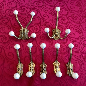 May include: Collection of antique brass coat hooks with white ceramic ball ends. The hooks are mounted on decorative brass backplates. The image shows a variety of hook designs, including double and single hooks.
