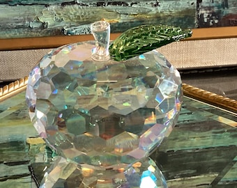 Op de afbeelding: Een helder, veelzijdig kristallen appelbeeldje met een reflecterend, geometrisch oppervlak dat het licht opvangt. De appel is bekroond met een kleine, transparante glazen steel en een enkel, gestructureerd, levendig groen glazen blad.