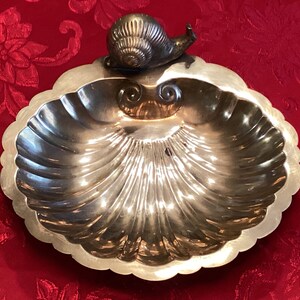 May include: Silver-coloured, shell-shaped dish with a scalloped edge. A small, detailed snail figurine sits atop the dish. The dish has a fluted design radiating from the centre. The background is a red floral pattern.