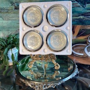 May include: Set of four clear glass coasters with silver trim, displayed on a gold metal stand. The coasters have a decorative, sunburst pattern.
