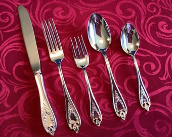 Op de afbeelding: Een set gepolijst zilver bestek, bestaande uit een mes, twee vorken en twee lepels. De handvatten hebben sierlijke details. Ze zijn gerangschikt op een rood, patroonrijk tafelkleed. Het mes staat links, gevolgd door vorken en lepels.