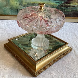 May include: A pink glass candy dish with a gold knob on the lid sits on a square, gold-framed mirror base.