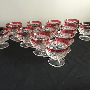 May include: Set of twelve clear glass cups with a red rim. The cups have a diamond pattern and are on a black surface.