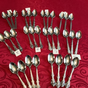 May include: A collection of 20 silver teaspoons with ornate designs. The spoons are arranged in rows on a red patterned surface.