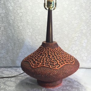 May include: A mid-century modern ceramic table lamp with a brown and orange textured base. The lamp has a wide, round base with a raised, textured design. The lamp is lit by a single bulb.