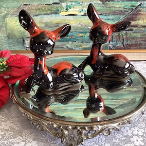 May include: Two black and orange ceramic deer figurines on a silver mirrored stand with an ornate design.