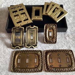 Pode incluir: Uma coleção de placas de interruptores de luz decorativas douradas com vários designs, incluindo designs ornamentados, simples e de dupla gang.