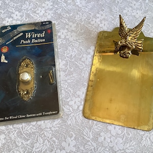 May include: A vintage brass doorbell push button with a decorative design, packaged in a clear plastic case. Beside it is a brass clipboard with an eagle emblem. The push button is labelled "Wired Push Button".