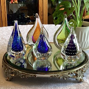 Puede incluir: Una colección de objetos de arte de vidrio en forma de lágrima en varios colores, incluyendo azul, naranja y verde, se exhibe en una bandeja con espejo con una base plateada ornamentada. Las piezas de vidrio tienen diseños de burbujas internas.