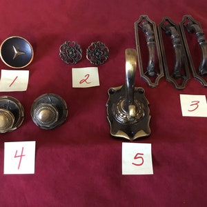 May include: Assortment of decorative hardware, including two ornate door knobs, two decorative floral accents, a black and gold round knob, and three ornate door pulls. The hardware is arranged on a red surface with numbered sticky notes.