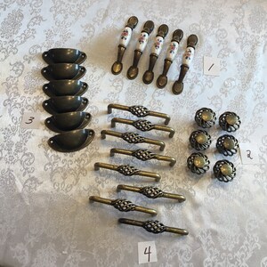 May include: Assortment of cabinet hardware in antique brass finish.  Includes 6 cup pulls, 6 knobs, 6 drawer pulls with a woven design, and 5 porcelain drawer pulls with floral designs.  Numbers 1, 2, 3, and 4 are written on pieces of paper.