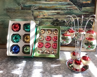 Könnte beinhalten: Eine Sammlung von Vintage-Weihnachtskugeln in verschiedenen Farben, darunter Rot, Grün, Gold und Blau. Einige Kugeln werden in ihren Originalverpackungen präsentiert, während andere an einem dekorativen Silberständer hängen. Die Kugeln weisen klassische Designs auf.