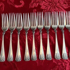 Puede incluir: Un conjunto de diez tenedores plateados con mangos ornamentados y púas. Los tenedores están dispuestos en una fila sobre un mantel rojo estampado. Cada tenedor tiene un diseño floral decorativo en el mango.