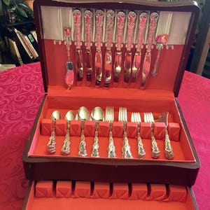 Puede incluir: Un cofre de cubiertos de madera con interior de terciopelo rojo, que muestra un juego completo de cubiertos de plata ornamentados. El juego incluye cuchillos, tenedores, cucharas y utensilios para servir, todos cuidadosamente dispuestos.