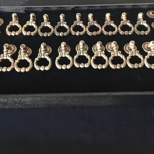 May include: An assortment of antique-style drawer pulls and decorative backplates in a display case. The pulls are ornate, with a ring-shaped handle and decorative detailing. The metal has a tarnished, aged appearance, suggesting vintage hardware.