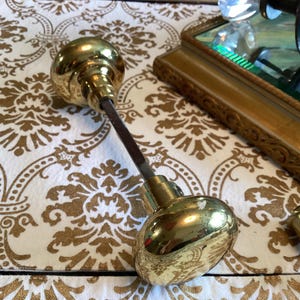 May include: A pair of polished brass door knobs connected by a square metal spindle. The round, smooth, reflective brass knobs are joined by a central rod, with an additional small brass cylindrical component positioned separately nearby.