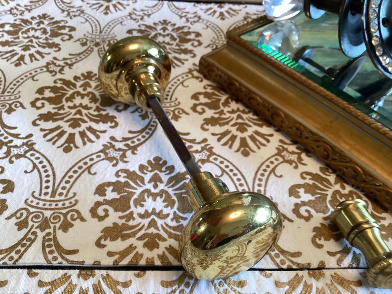 May include: A pair of polished brass door knobs connected by a square metal spindle. The round, smooth, reflective brass knobs are joined by a central rod, with an additional small brass cylindrical component positioned separately nearby.