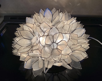 Op de afbeelding: Een bolvormige lamp opgebouwd uit talloze doorschijnende, gebroken witte, bloembladachtige panelen die in een gelaagde, bloemvormige structuur zijn gerangschikt. Elk paneel is omlijnd met een dunne, donkere rand voor een geometrisch effect.